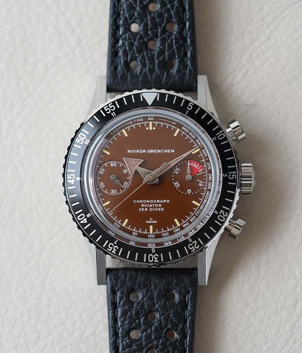 A Nivada Grenchen Chronomaster Broad Arrow watch with a black racing strap and stainless steel case, featuring a matte black dial with cream colored luminous indexes and shiny silver hands.